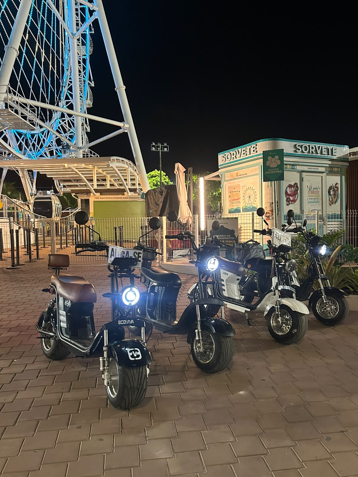 Moto elétrica preta da VoltGo estacionada à noite na orla, com palmeiras iluminadas em verde ao fundo
